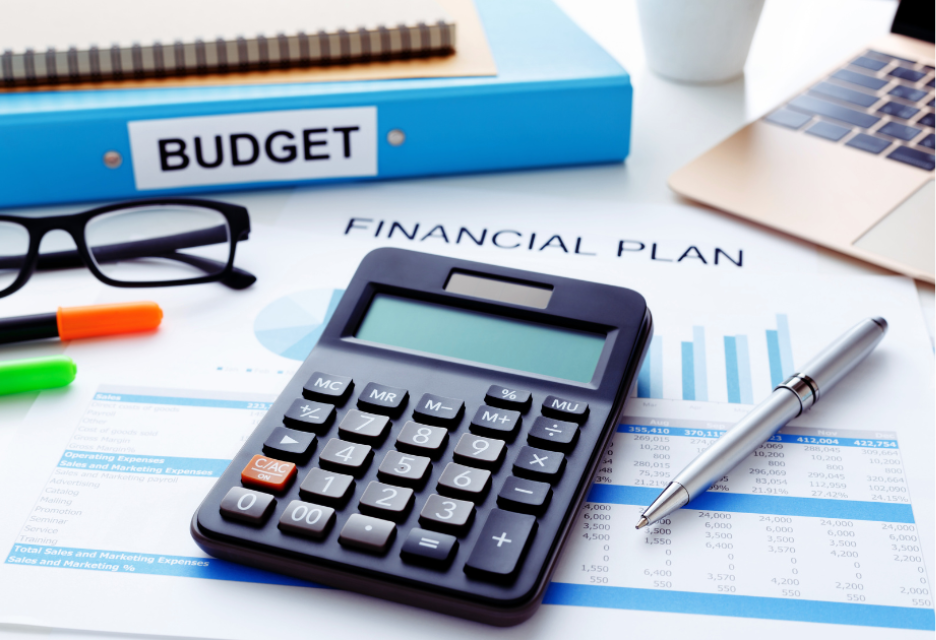 A financial planning workspace featuring a calculator, budget binder, financial plan documents, charts, a laptop, glasses, and pens on a desk.
